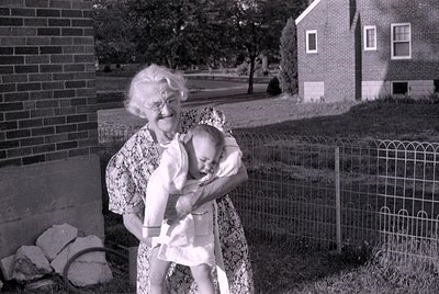 A black and white photograph of an older woman with white hair and glasses holding a baby. They are outdoors in a yard with a...