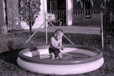A black and white photograph of a toddler wearing dark swim trunks standing in a small, inflatable circular pool. A wire fenc...