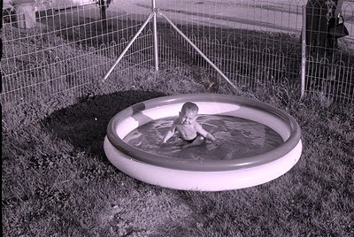 A small child sits in a circular inflatable wading pool placed on a grassy lawn. A wire fence and metal support poles are vis...