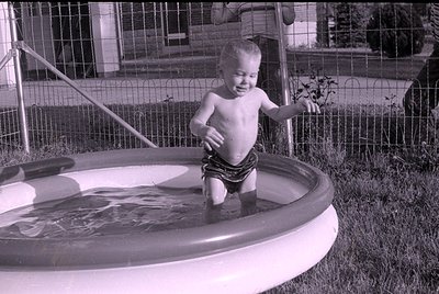 A black and white photograph of a shirtless young child standing in a small, inflatable swimming pool. A wire fence and a bui...