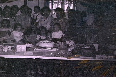 A group of children and adults stand behind a table featuring a frosted cake and various dishes. The image is a dark, purple-...