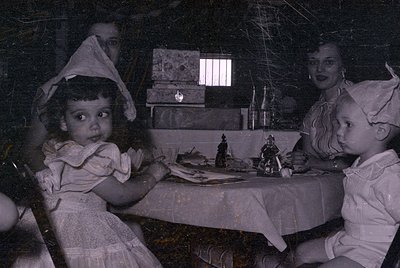 A black and white photograph showing two young children in white dresses and bonnets sitting at a table with a white tableclo...