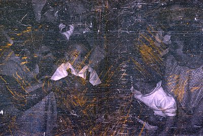 A heavily damaged photograph showing fragmented figures of people. The image is obscured by deep scratches, chemical staining...