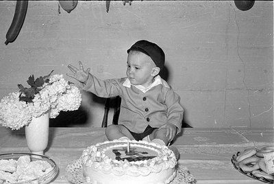 A black and white photograph of a young child wearing a dark cap and a light-colored button-up jacket. The child is seated at...
