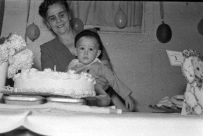 A black and white photograph of a woman and a young child behind a table with a frosted cake. A small card in the background ...