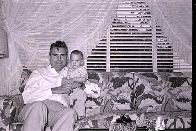 A man in a white button-down shirt and light-colored trousers sits on a patterned sofa holding a baby. Behind them are white ...