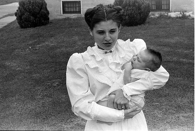A black and white photograph, oriented upside down, showing a woman holding an infant. The baby is wearing a white, ruffled g...