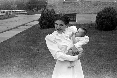 A black and white photograph of a woman in a white, ruffled long-sleeved dress holding an infant. They are outdoors on a lawn...
