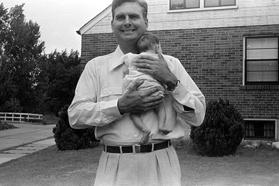 A black and white photograph of a man standing in front of a brick building, holding an infant. The man is wearing a light-co...
