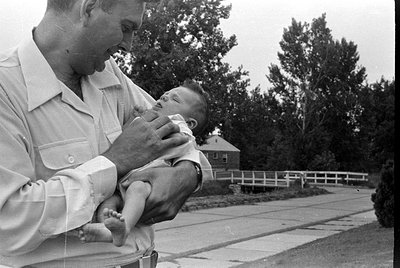 A black and white photograph of a man in a light-colored button-down shirt holding an infant. The scene is outdoors on a pave...