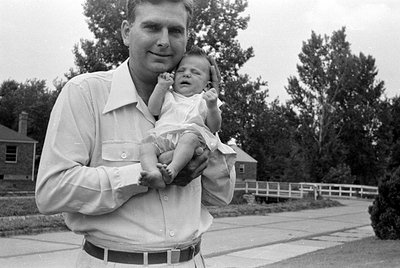 A black and white photograph of a man in a light-colored button-down shirt holding an infant. The baby is wearing a white dre...