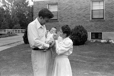 A man in a light-colored button-down shirt and trousers holds an infant. A woman in a white dress stands beside them, looking...