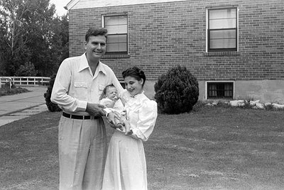 A black and white photograph of a man, a woman, and an infant standing in front of a brick building. The man wears light-colo...