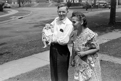 A black and white photograph of a man, a woman, and an infant standing outdoors on a sidewalk. The man wears a light-colored ...