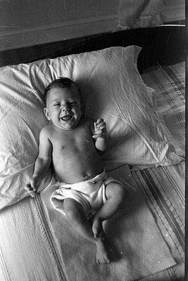A black and white photograph of an infant lying on a bed. The baby is wearing a diaper and is positioned on a white pillow an...