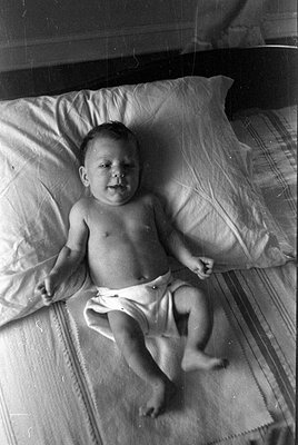 A black and white photograph of an infant lying on a bed. The baby is wearing a diaper and is positioned on a white pillow an...