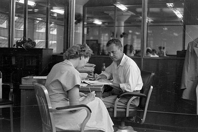 A man and a woman sit at a desk in an office with large glass partitions. The woman is writing on a piece of paper while the ...