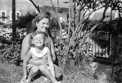 A black and white photograph of a woman and a young girl sitting outdoors. The woman is positioned behind the girl, with her ...