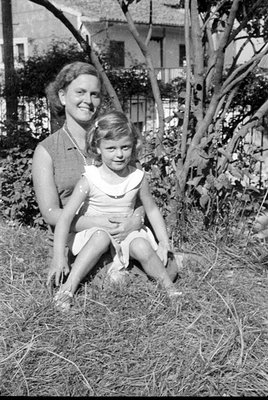 A black and white photograph of a woman and a young girl lying in tall grass. The woman wears a dark sleeveless dress and a p...