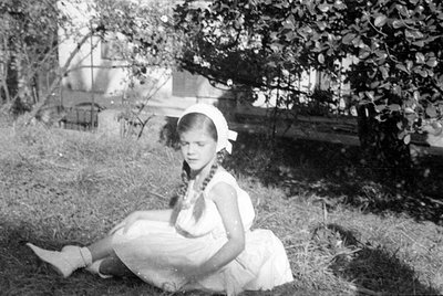 A young girl with braided hair and a white headpiece sits on a grassy slope. She is wearing a light-colored sleeveless dress ...