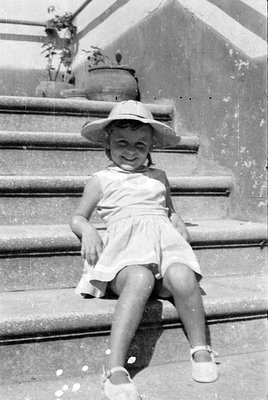 A young girl wearing a light-colored sleeveless dress and a wide-brimmed sun hat sits on stone steps. She is wearing white st...