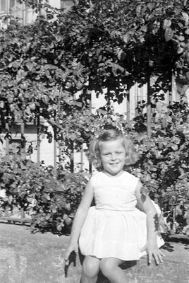 A young girl in a light-colored, sleeveless dress sits outdoors in front of dense foliage and a white fence. She is smiling a...