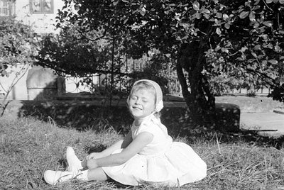A young girl in a white dress and white shoes sits on a grassy lawn. She is wearing a light-colored headband and has her eyes...