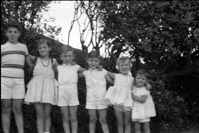 Six children stand in a row outdoors against a backdrop of dense foliage. They are wearing light-colored summer clothing, inc...