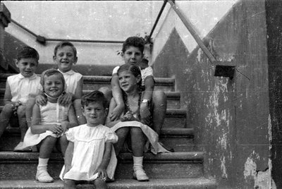 Six children are posed on a set of concrete stairs. They are wearing light-colored summer clothing, including dresses and sho...