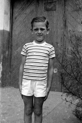A full-length black and white portrait of a young boy standing outdoors. He is wearing a short-sleeved horizontally striped t...