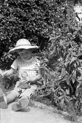 A black and white photograph of a child wearing a wide-brimmed sun hat and a light-colored patterned shirt. The child is sitt...