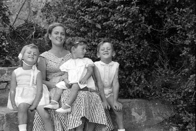 A black and white photograph of a woman and three young children sitting on a stone ledge. The woman wears a striped dress, a...