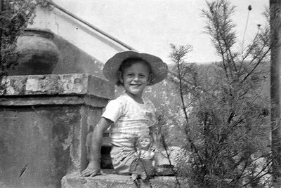 A black and white photograph of a young child wearing a wide-brimmed hat and a light-colored t-shirt. The child is leaning ag...