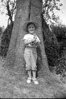 A black and white photograph of a child standing in front of a large tree trunk. The child is wearing a wide-brimmed hat, a l...