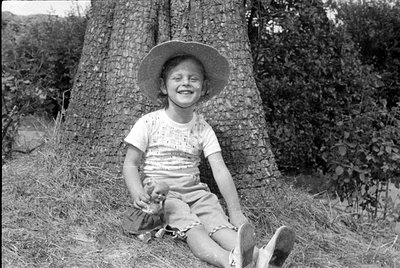 A black and white photograph of a smiling child wearing a wide-brimmed sun hat and light-colored shorts. The child is sitting...