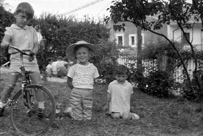 Three children in a grassy yard. One child stands with a bicycle on the left, another wears a wide-brimmed hat in the center,...