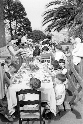A black and white photograph of a group of adults and children gathered around a long table set for a meal outdoors. A woman ...