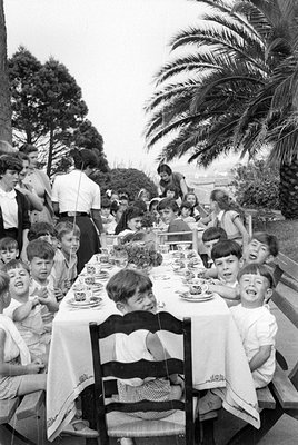 A group of children and adults are gathered around a long table covered with a white cloth in an outdoor setting. The table i...