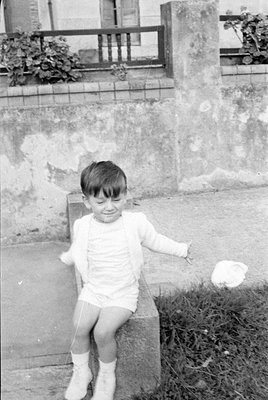 A young child wearing a white short-sleeved outfit, a white cardigan, and white socks sits on a concrete block. The backgroun...