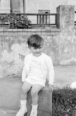 A black and white photograph of a young boy sitting on a stone block. He is wearing a light-colored cardigan, matching shorts...