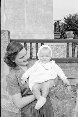 A black and white photograph of a woman holding a baby. The baby wears a white bonnet and a white dress. They are positioned ...