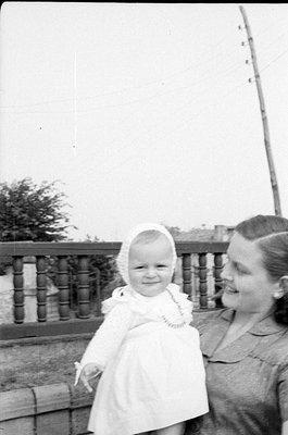 A black and white photograph of a woman and a small child. The child wears a white dress and a matching bonnet, while the wom...