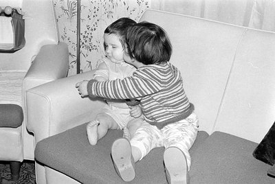 Black and white photograph of two young children sitting on a textured sofa. One child wears a striped long-sleeved shirt and...