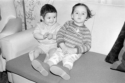 Two young children sitting on a light-colored sofa with a blue cushion. The child on the left wears a light-colored short-sle...