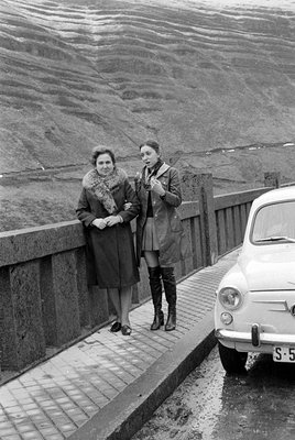 Two women stand on a concrete bridge walkway next to a light-colored small car. One woman wears a dark coat with a fur collar...