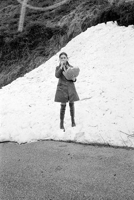 A black and white photograph of a woman standing in front of a large pile of snow. She is wearing a dark knee-length coat and...