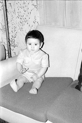 A black and white photograph of an infant lying on a patterned surface next to a grey upholstered piece of furniture. The bab...