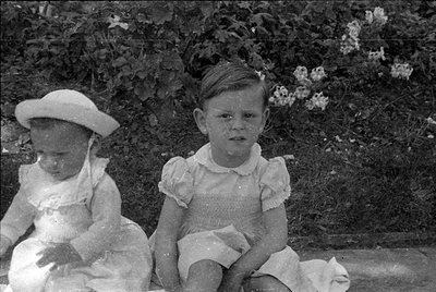 Two young children sitting outdoors in front of a hedge with small white flowers. Both wear light-colored, ruffled dresses; t...