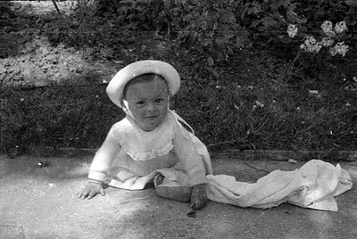 A black and white photograph of a young child sitting on a concrete surface outdoors. The child wears a light-colored hat and...