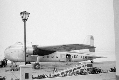 A side view of a propeller airplane with the registration EC-AEH on its fuselage. Two people stand near the open cabin door o...